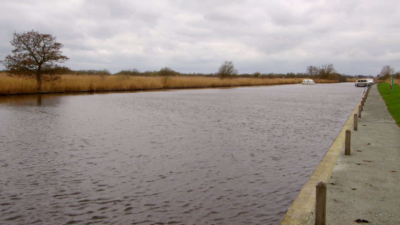 Fremskyndet optagelse, der kigger op ad floden Bure med en hvid Norfolk Broads-cruiserbåd, der passerer fortøjede både