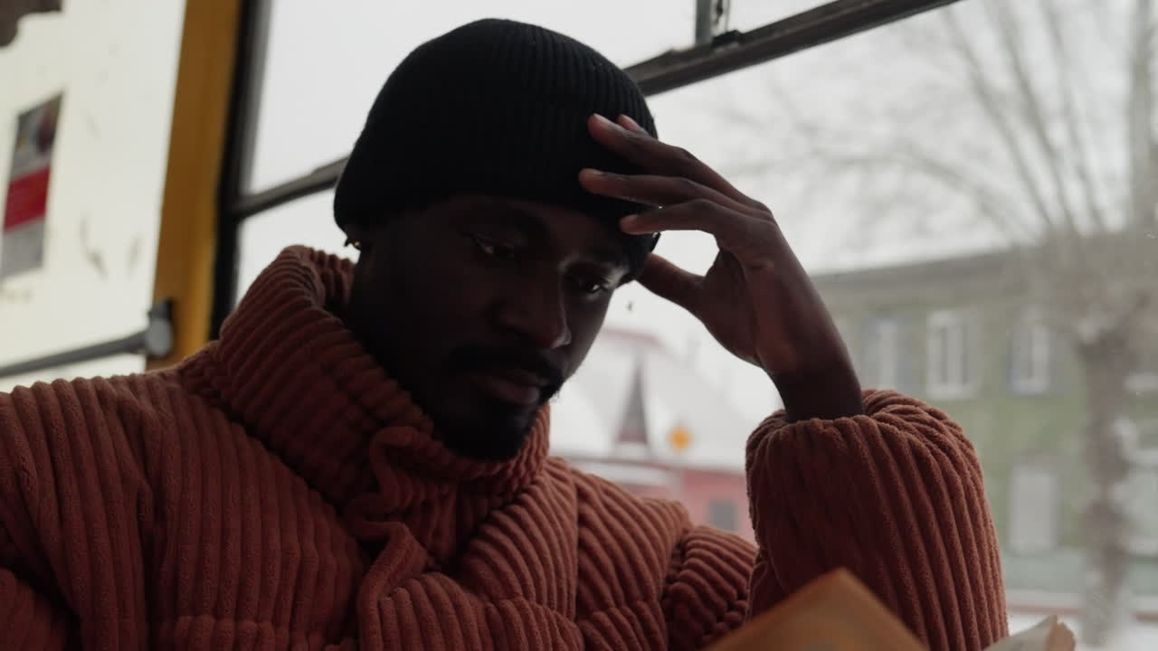 Black man on bus window pensive, wearing chunky knit sweater and beanie, gazing at snowy street, city buildings blurred outside, warm tonal palette, soft natural light, quiet introspective mood