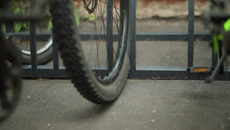 close-up of bicycle wheel parked in metal bike rack, capturing spokes and frame details, background features additional bicycles and blurred greenery