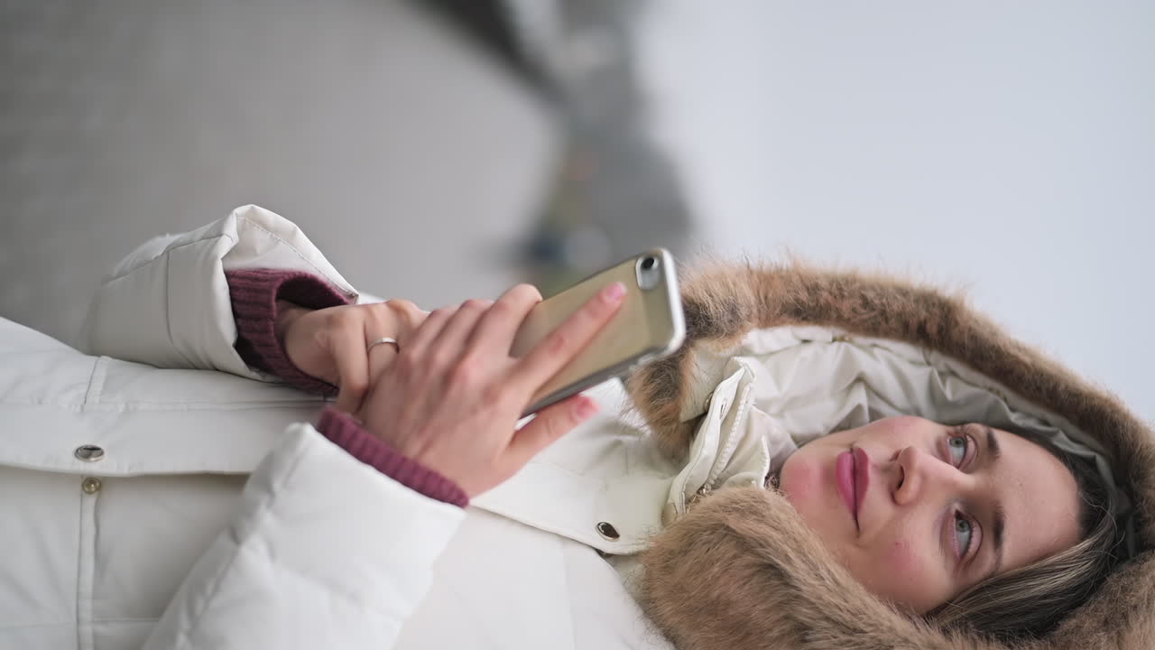 A woman in a warm, white coat stands in a park during a cool day. She focuses on her phone, absorbing the quiet surroundings and embracing the serene atmosphere