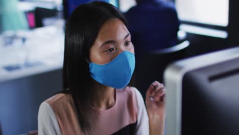 asian woman wearing face mask using computer while sitting on her desk at modern office