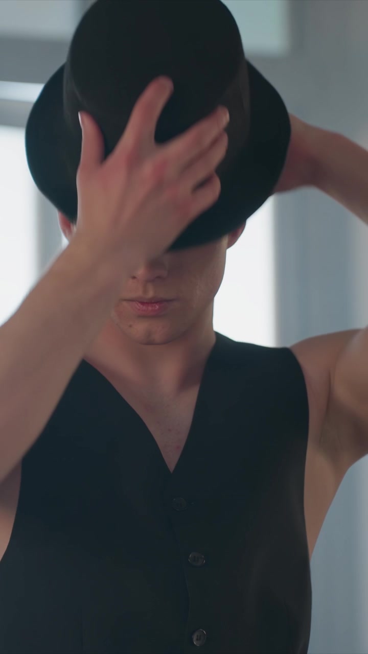 Giovane uomo si sistema ripetutamente il cilindro nero e si liscia il... L'uomo indossa un gilet nero senza maniche di colore scuro. La scena presenta fonti di luce intensa e interni moderni.