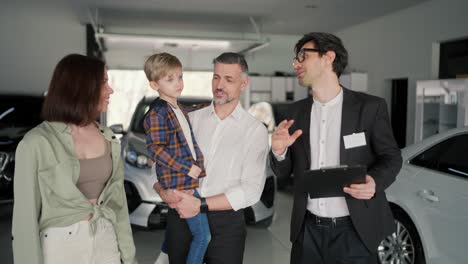 Close-up-a-confident-male-brunette-assistant-with-glasses-in-a-business-suit-with-a-tablet-in-his-hands-tells-the-family-about-what-cars-are-in-the-car-dealership-and-about-modern-car-models