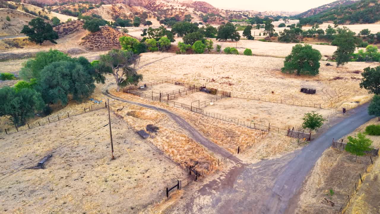 An aerial drone view of empty livestock corrals on a rustic ranch in California. The metal pens sit in a dry, golden valley, ready for cattle or horses