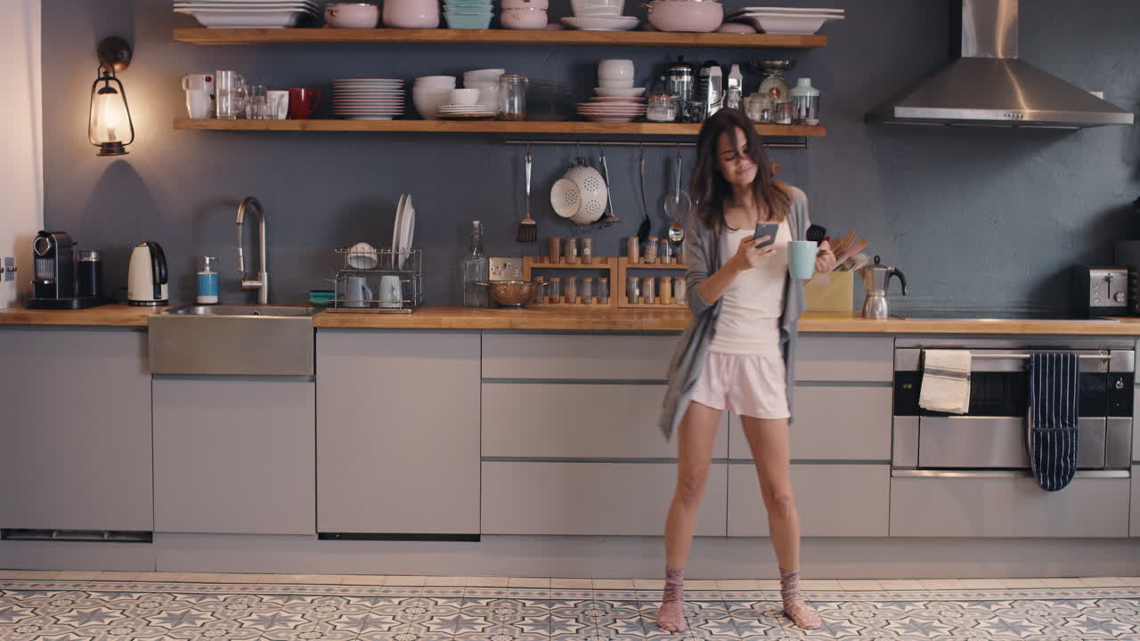 mujer feliz bailando en el bucle de la cocina chica divertida usando teléfono inteligente divirtiéndose bailando bebiendo café usando pijama en casa rutina matutina tonta
