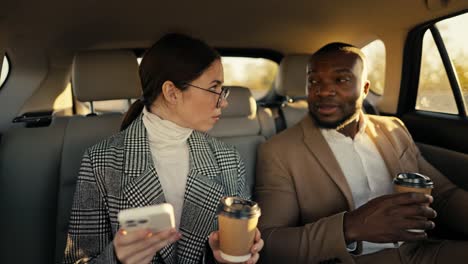 A-confident-brunette-businesswoman-in-round-glasses-with-a-cup-of-coffee-in-her-hands-and-a-smartphone-communicates-with-her-colleague-a-man-with-Black-skin-and-a-brown-jacket-during-his-business-trip-in-the-cabin-of-a-modern-car-outside-the-city