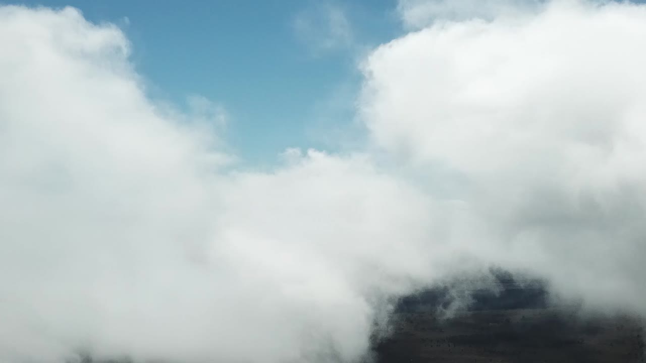 Billowing low-altitude clouds drift over dark volcanic slopes, partially revealing the rugged terrain beneath a bright, cloud-dappled blue sky.