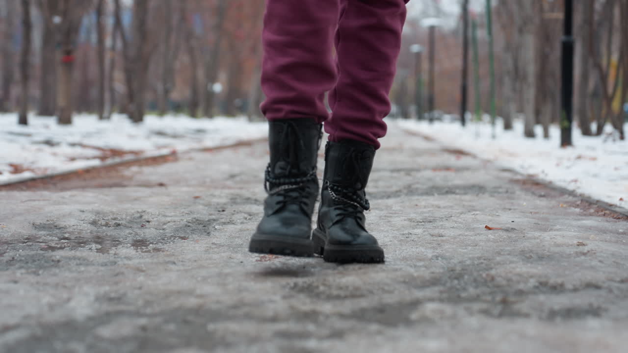 primer plano de las piernas en pantalones de color marrón y botas de diseño negras pisando un camino cubierto de nieve helada, el fondo borroso presenta árboles desnudos y farola