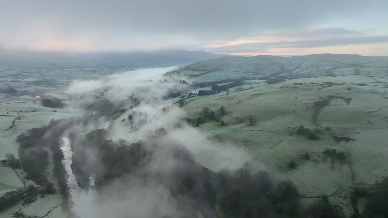 el campo de patchwork nublado en la ladera de la colina con el río envuelto en la niebla y el resplandor del amanecer en el horizonte al amanecer en invierno