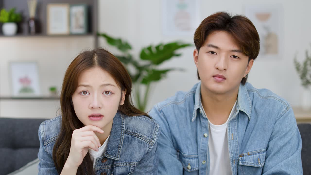 คู่รักชาวเอเชียวัยหนุ่มสาวแสดงอาการตกใจและหงุดหงิดขณะดูโทรทัศน์ในห้องนั่งเล่น