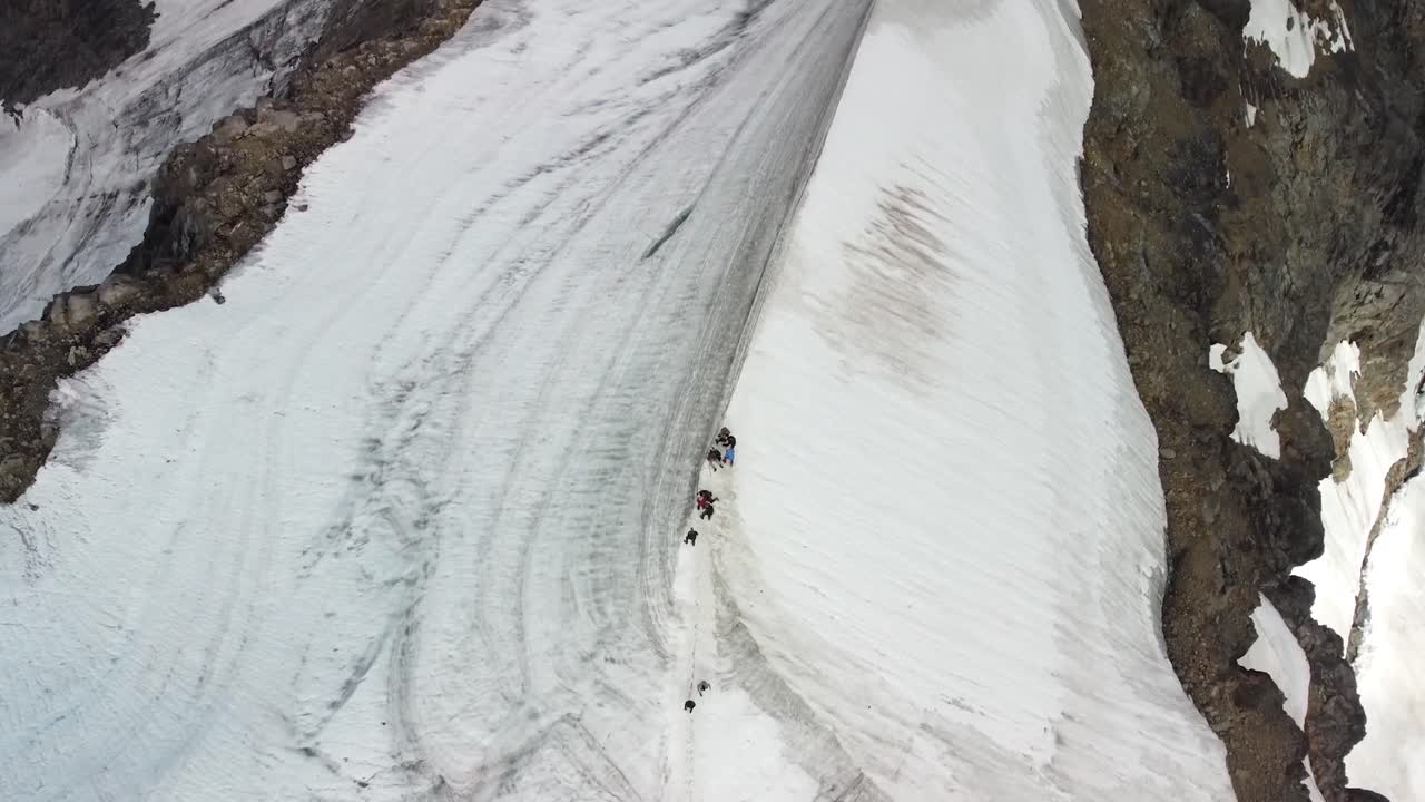 Top down aerial drone footage of group of people walking and hiking on tall white snow covered Kebnekaise mountain edge or cliff during a cloudy day. Mountain itself is black colored and volcanic.