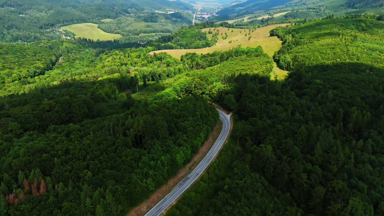 Mountain valley with road between green hills.. Aerial view of a narrow road curving through forested hills. The valley opens into fields and distant mountains