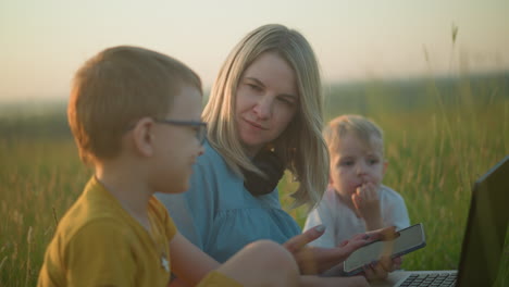 a joyful mother in a blue gown multitasks on a laptop and phone in a grassy field, engaging with her son in yellow, who laughs while her other son in white sits quietly beside them