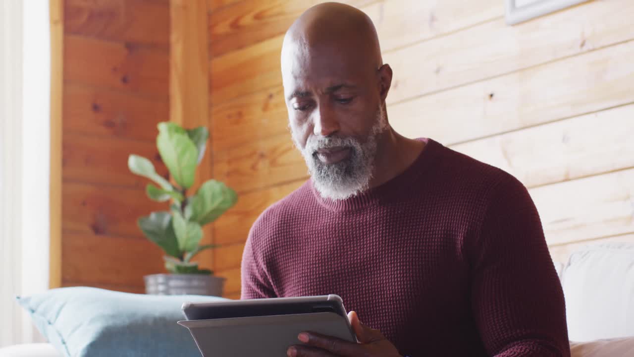 Happy senior african american man spending time in log cabin, sitting on sofa and using tablet