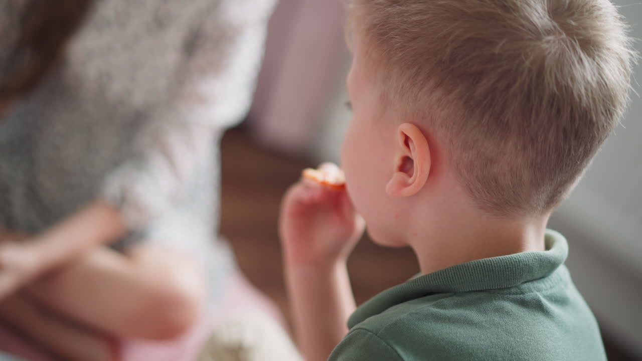Close up of little boy in green shirt holding slice of pizza, preparing to take bite, with blurred view of another child sitting nearby in soft natural light