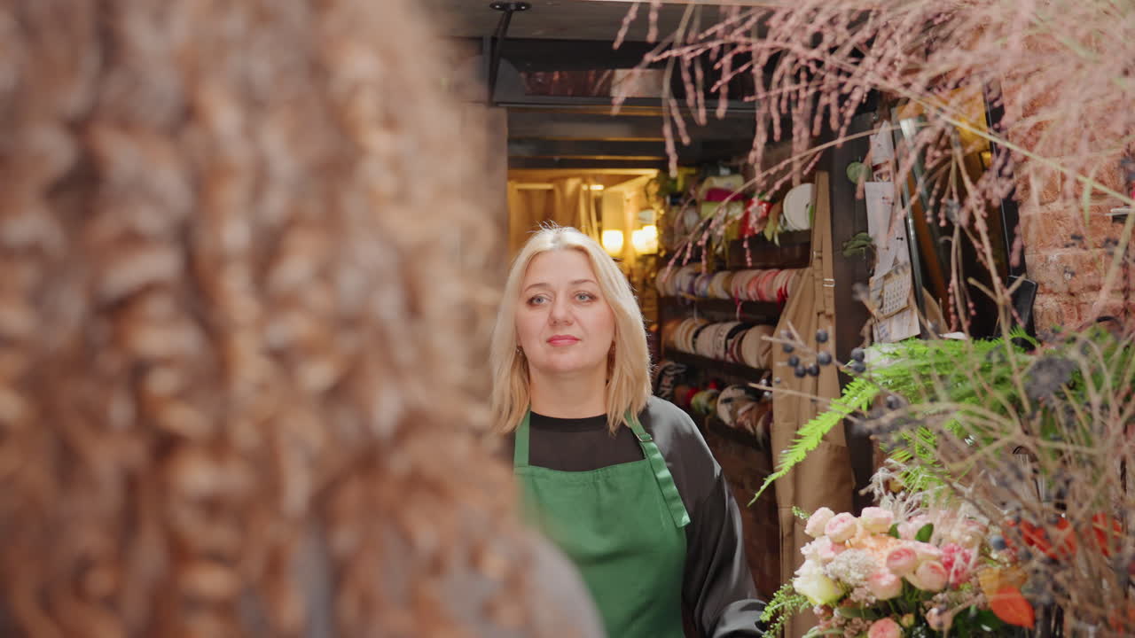 Assistentin der Floristin überreicht Blumenstrauß an lockenhaarige Kundin an der Theke, lächelnde Assistentin mit verpackten Pfingstrosen und Grünzeug, Boutique-Regale mit Bändern und Vasen, lockerer Austausch