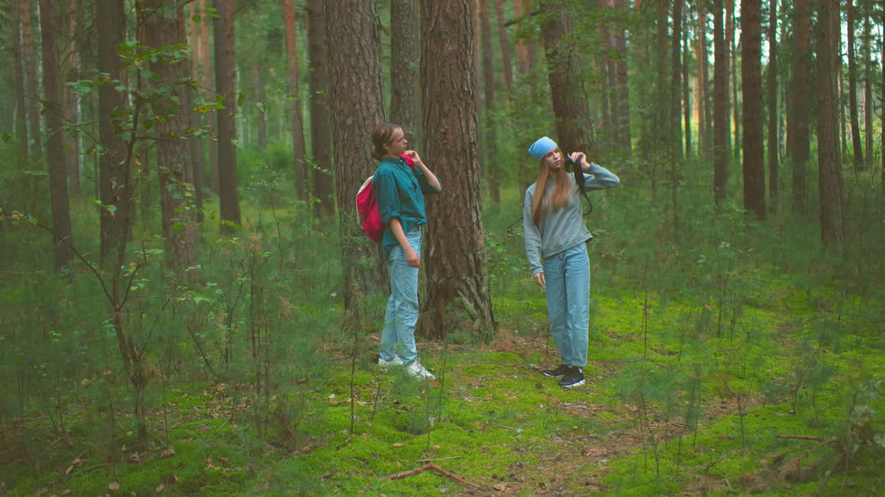 여성들은 평화로운 숲에서 가방을 가져오고, 높은 나무와 울창한 초록색으로 둘러싸인 산책을 계속할 준비를 하고, 가방을 어깨에 매달고, 멀리 고 있습니다.