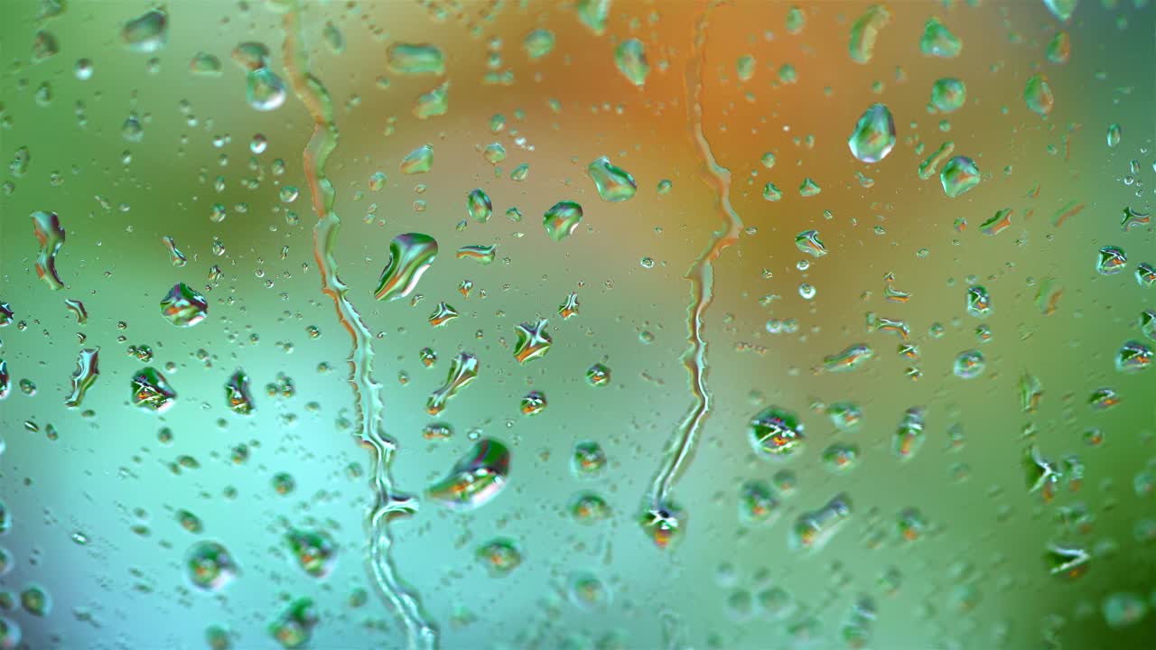 Raindrops on blur colorful background. Large drops flowing on glass window slowly in rainy weather. Close-up.