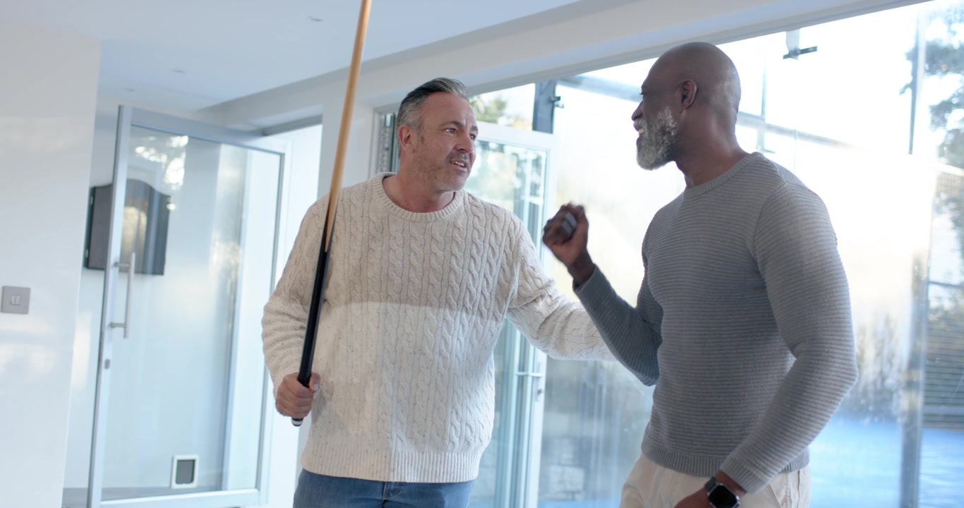 Three male friends at pool table watching cream-sweater man striking cue ball to pocket balls