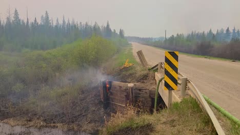 Overturned-car-on-roadside-engulfed-in-flames-with-smoke,-in-daylight,-conveying-urgency-and-disaster,-rural-setting