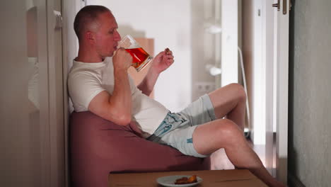 business man seated casually in hallway eats goat meat while drinking beer from large glass cup with his reflection visible on door, relaxing after long workday, surrounded by simple home interior