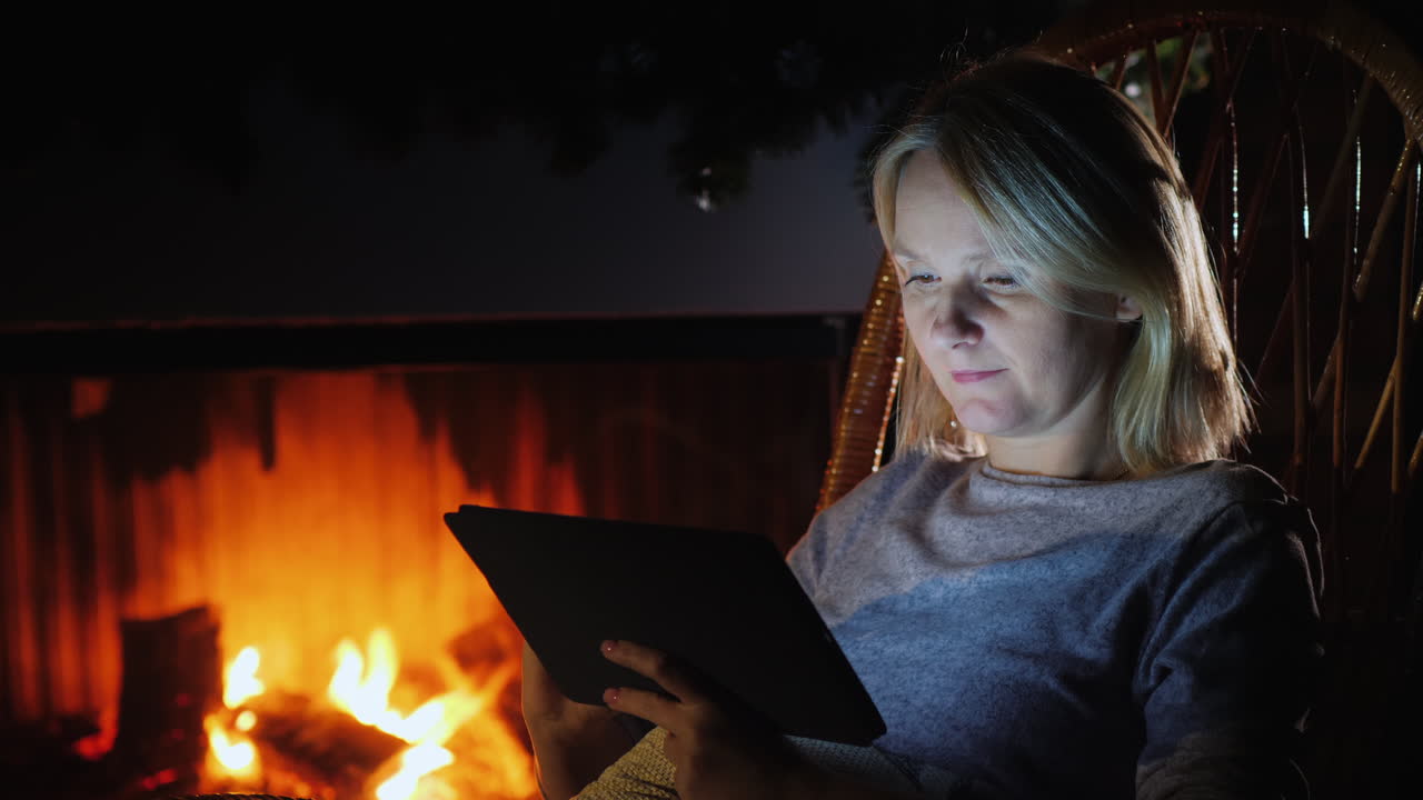 una joven usa una tableta digital en una casa acogedora cerca de la chimenea donde arde la llama