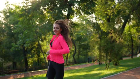 Young-woman-with-long-hair-in-bright-pink-jacket-running-in-the-sunny-city-park-and-looking-in-the-camera.-Steadicam-stabilized