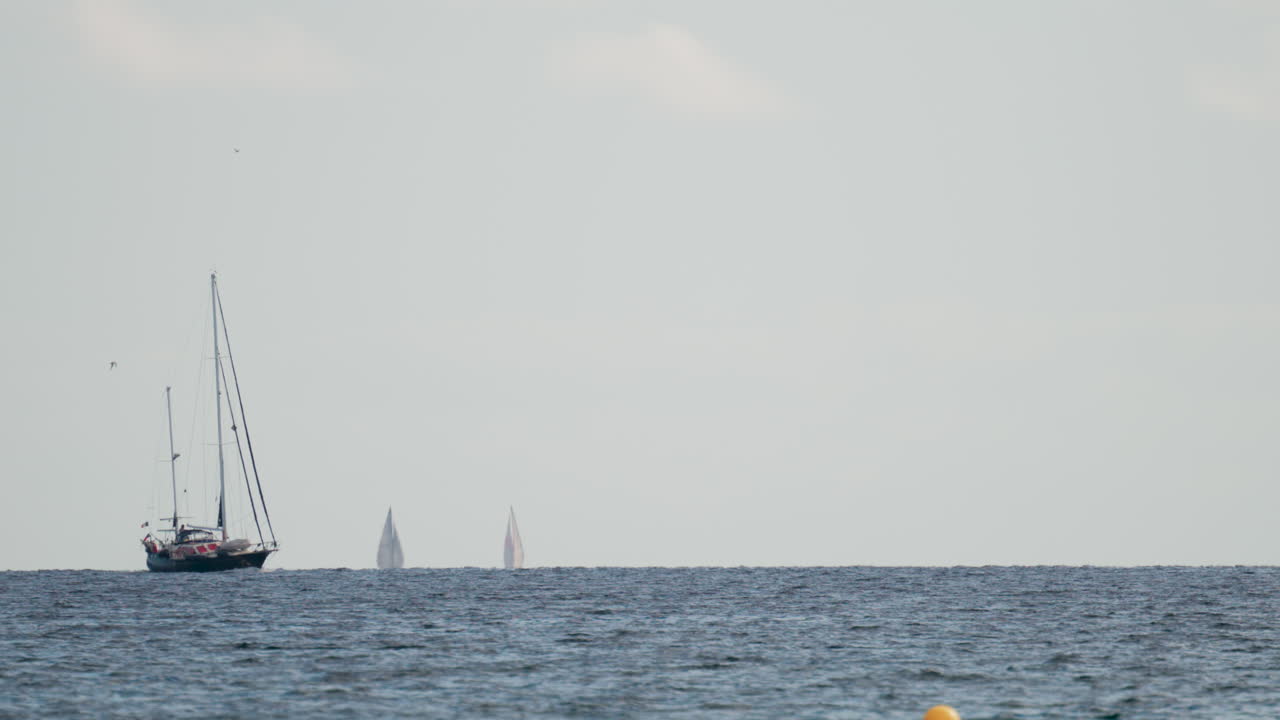 Wide shot of two sailboats navigating the calm blue sea under a pale sky