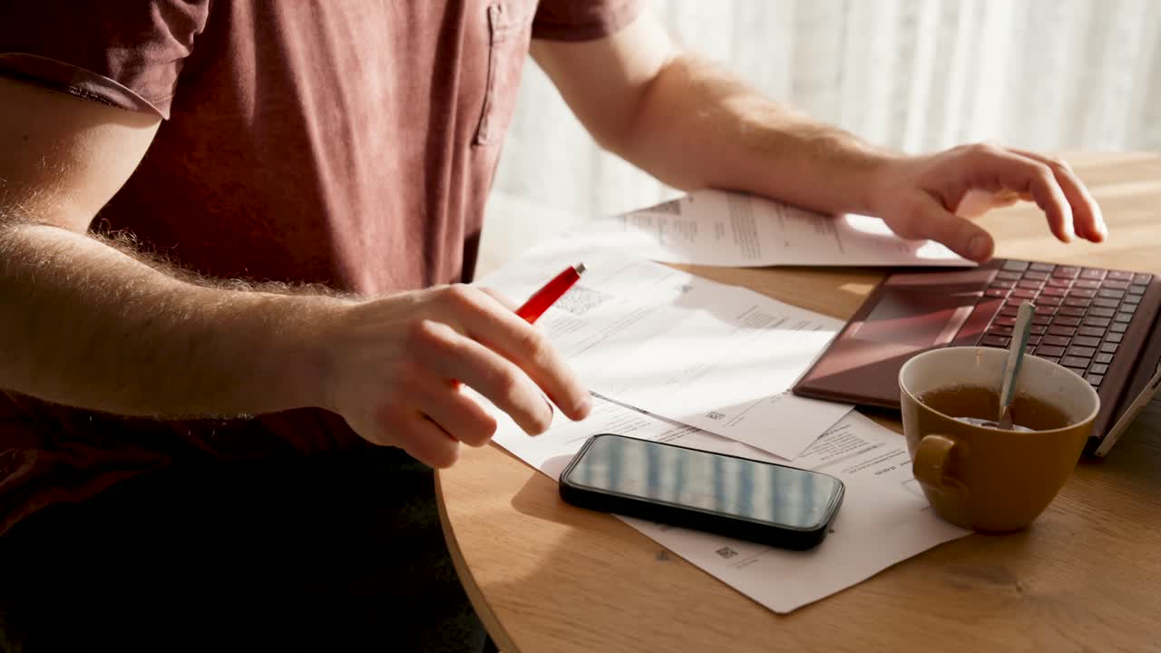 a man sits at a table in the sunshine and types something on his cell phone. lots of paper on the table. he pays his bills