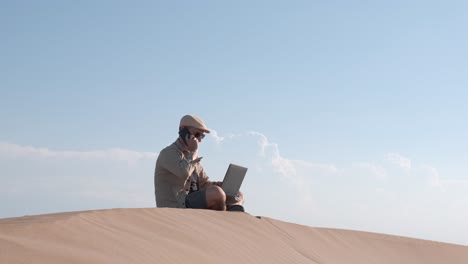 male traveler sitting on sandy dune using laptop. digital nomad or remote worker concept