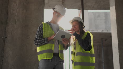 Dos-Ingenieros,-Un-Hombre-Y-Una-Mujer-Con-Cascos-Blancos-Y-Una-Tableta-En-El-Sitio-De-Construcción,-Condenan-El-Plan-De-Construcción-Del-Edificio