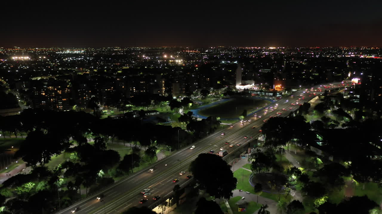 An aerial view after dark, showcasing a bustling highway and park in Buenos Aires, where traffic flows amidst the city's illuminated buildings