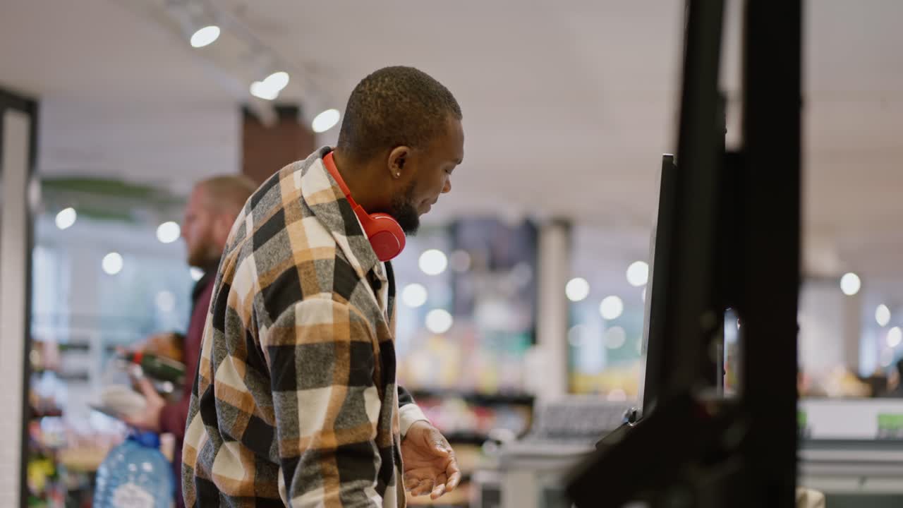 Side view of a man with Black skin color and a beard in a plaid shirt and red wireless headphones scans the goods he needs at a self-checkout checkout in a modern spacious light grocery store