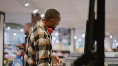 Side-view-of-a-man-with-Black-skin-color-and-a-beard-in-a-plaid-shirt-and-red-wireless-headphones-scans-the-goods-he-needs-at-a-self-checkout-checkout-in-a-modern-spacious-light-grocery-store