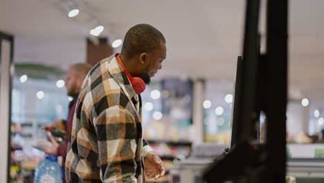 side view of a man with black skin color and a beard in a plaid shirt and red wireless headphones scans the goods he needs at a self-checkout checkout in a modern spacious light grocery store