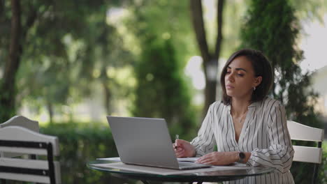 student woman brunette hispanic ethnos studying remotely via the internet while sitting in a summer cafe with a laptop and writing down a pen and a notebook while taking notes on the webinar