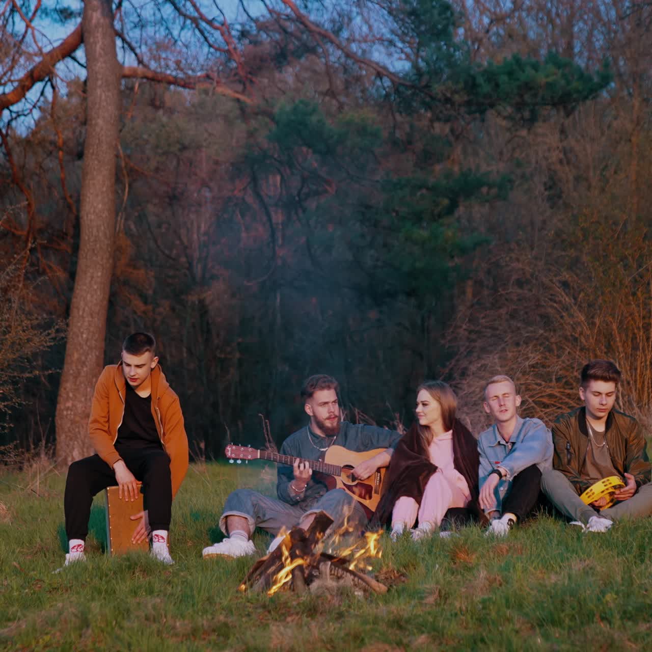 Evening near the fire outdoors. Group of friends sitting near the campfire in the forest. Young friends singing to the guitar in nature