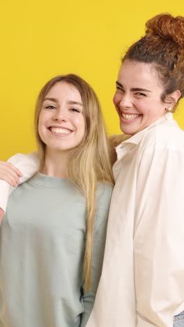 two young women smiling and embracing on yellow background