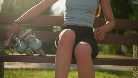 partial view of lady dropping cyan roller skates on wooden bench while sitting, with abdomen slightly exposed under sunlight, background features greenery and sun shining from behind