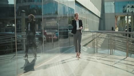 professional banker walking confidently with coffee in hand on phone call near glass wall and iron railing, reflecting urban scenery and cars, showcasing business lifestyle in modern environment