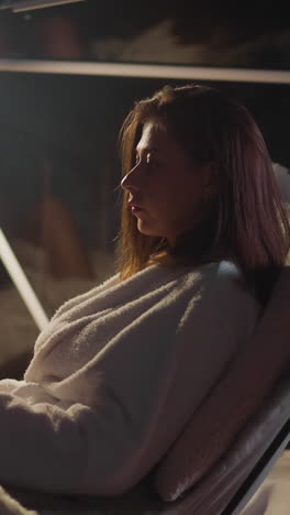 elegant woman dressed in bathrobe puts leg over leg sitting at table. serious lady rests by panoramic window with cup of hot drink enjoying pleasant evening