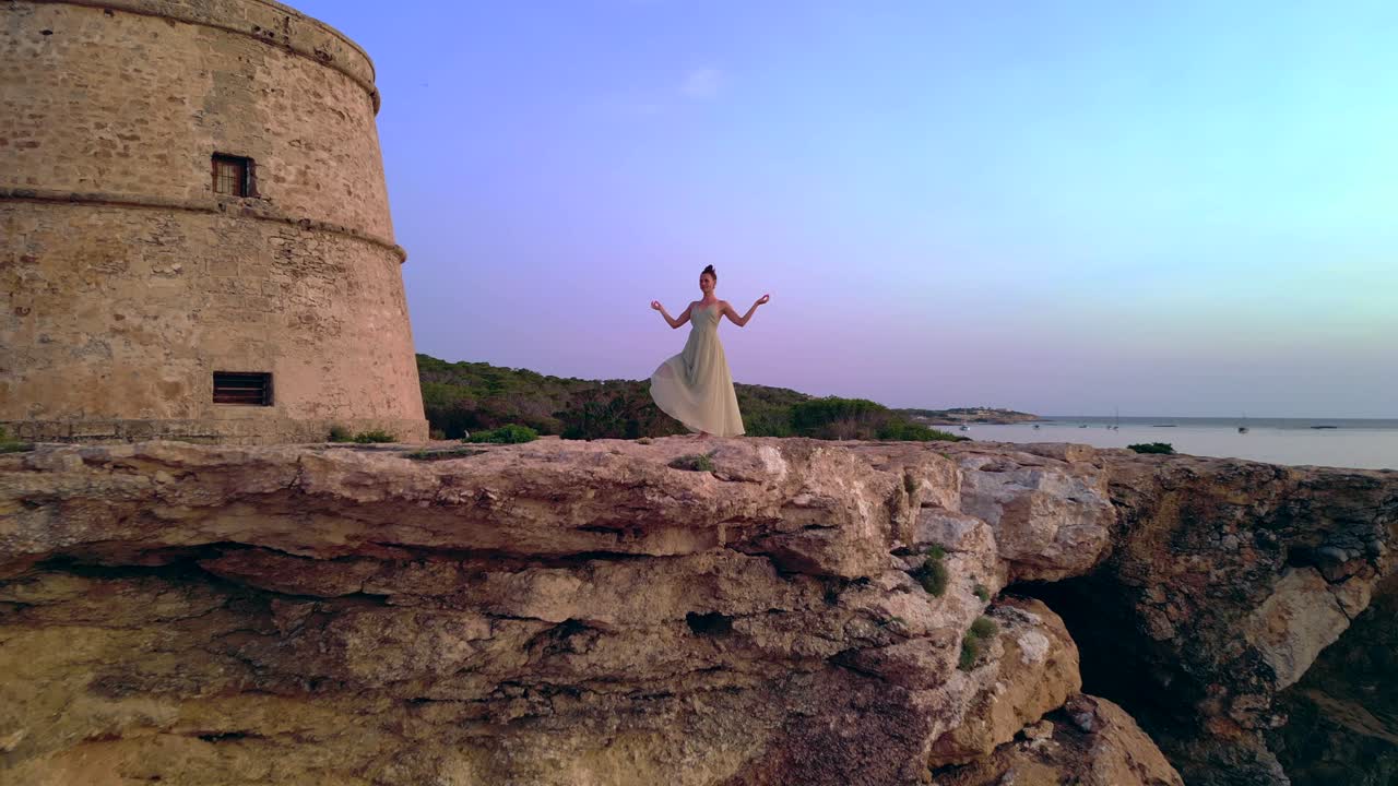 Barefoot hippie girl practicing yoga at sunset near Torre des Carregador, Ibiza, Spain. speed ramp hyper motion time lapse. overflight flyover drone