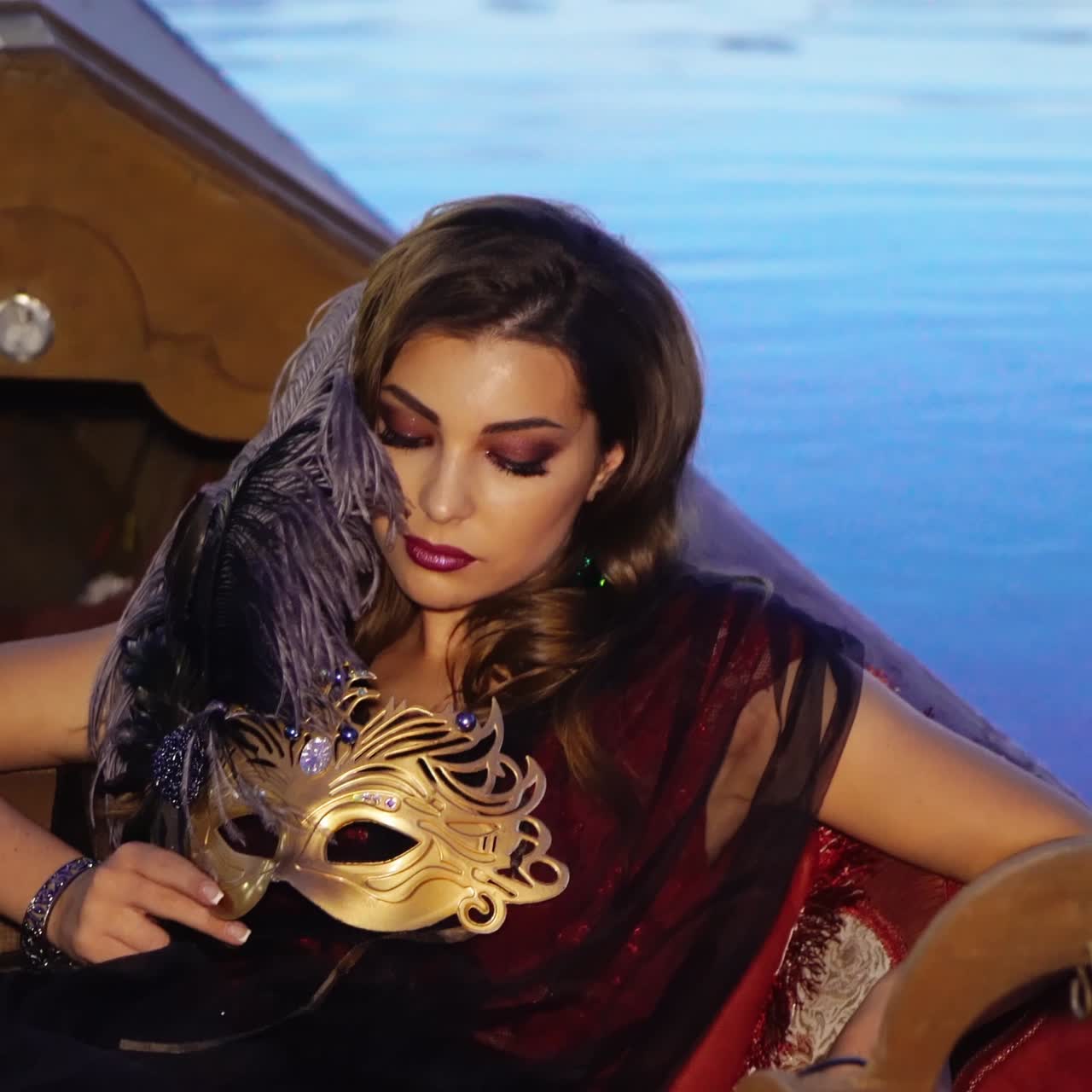 A gorgeous model in a dress is sitting in the gondola and holding a mask in golden colour with precious stones and a purple feather in her hand on the background of water in the evening.