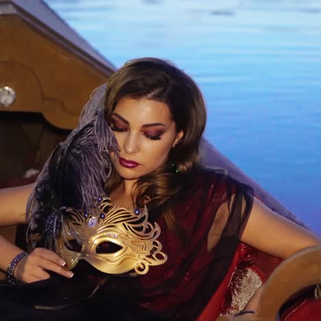 a gorgeous model in a dress is sitting in the gondola and holding a mask in golden colour with precious stones and a purple feather in her hand on the background of water in the evening.