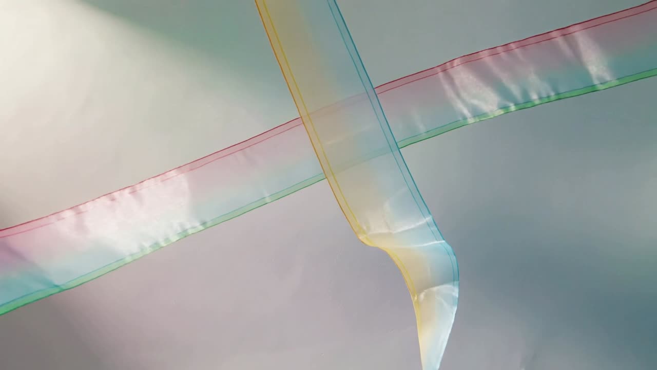 Descending yellow ribbon meeting pink teal ribbon, twisting and forming X in studio lit upper left