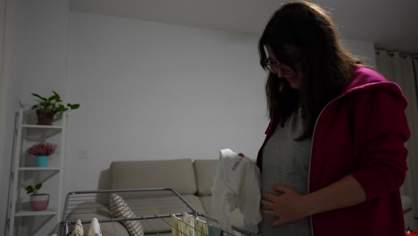 pregnant woman smiles while holding a baby onesie near her belly, standing next to a drying rack in living room. medium shot