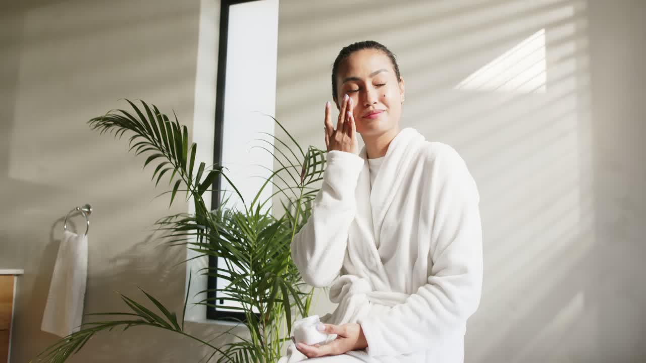 Mid adult Asian woman unscrewing white cream jar lid and smoothing under-eye cream in spa bathroom