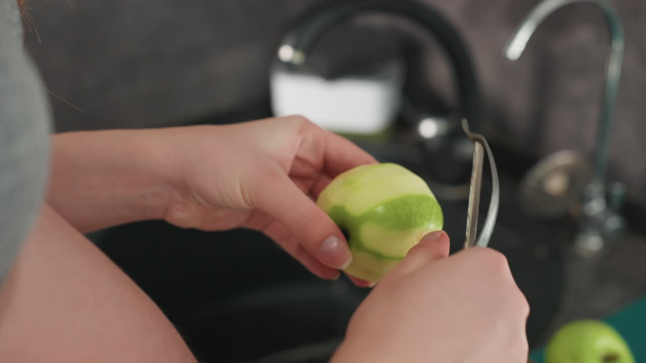 Primer plano de mujer embarazada pelando una manzana junto al fregadero, toma de cerca de la mano mostrando el pelador deslizándose sobre la piel verde, enjuague y lavabo al fondo, textura táctil y movimiento preciso, merienda prenatal consciente.