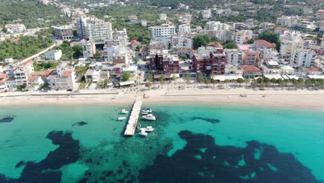 Drone-view-in-Albania-flying-over-a-beach-with-crystal-clear-blue-water-ocean,-buildings-on-the-harbor-and-a-wooden-walkway-on-a-sunny-day