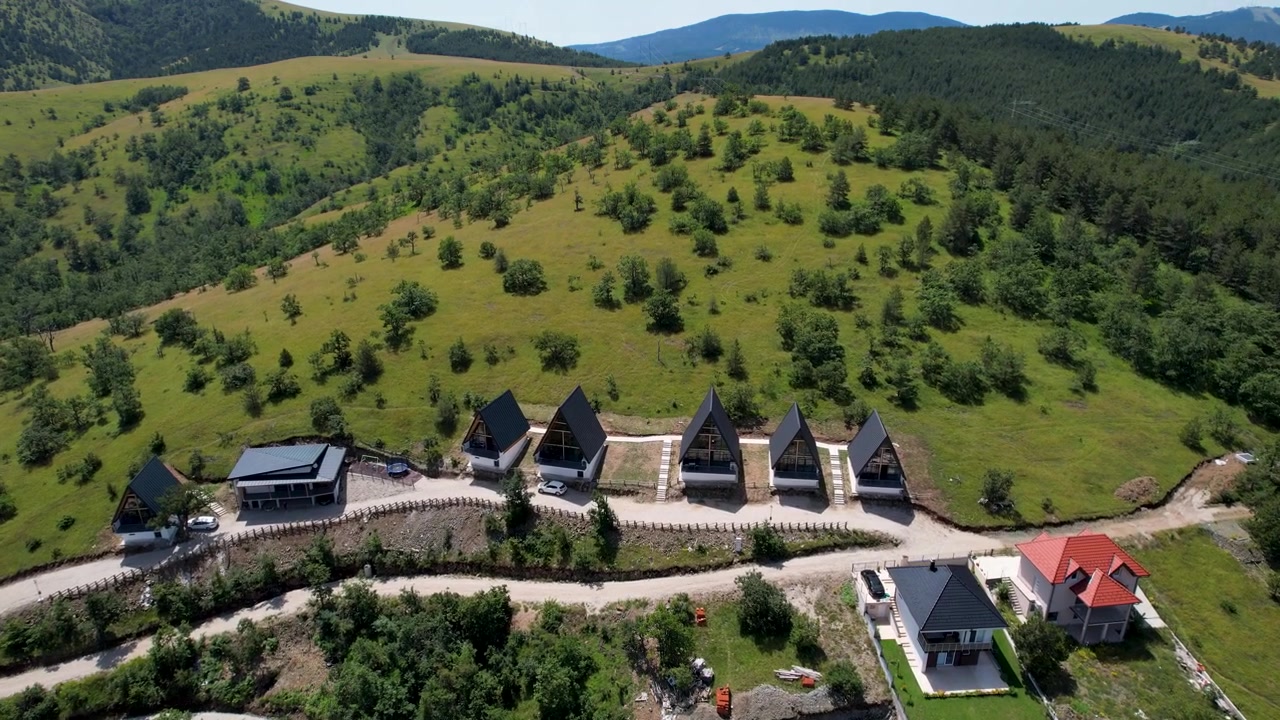 Dron przelatuje nad rzędem górskich chatek z ciemnymi dachami, poniżej otwartych, trawiastych wzgórz i rozrzuconych drzew w jasnym świetle dziennym w Zlatiborze, w Serbii, w słoneczny dzień